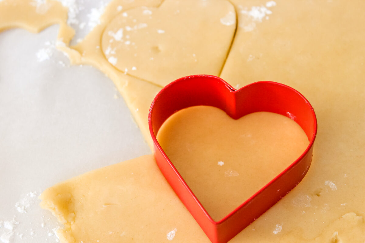 Sugar cookies being cut out with a heart shaped cookie cutter.